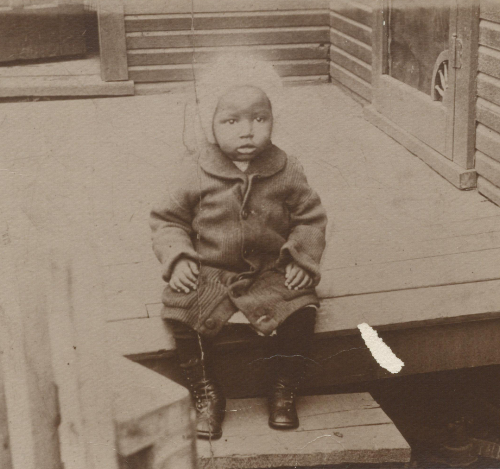 Strayhorn, a toddler, sits in coat and cap on edge of wooden porch and house.